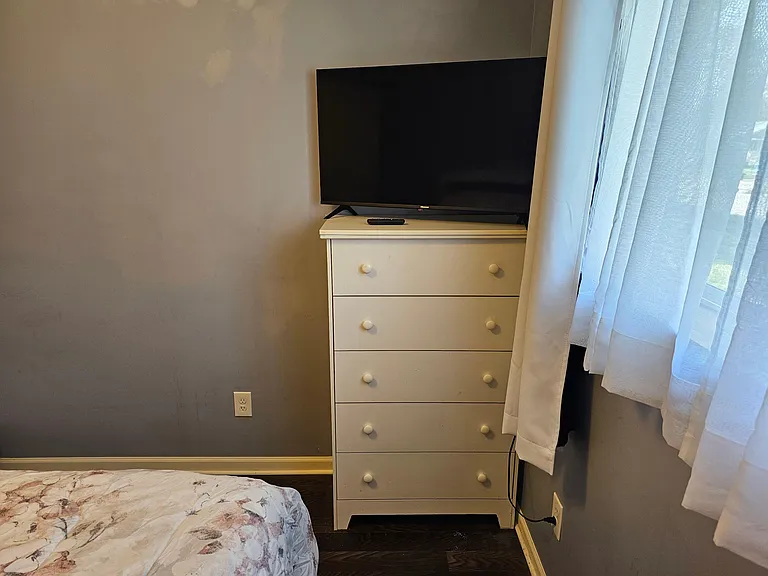 Bedroom area with a dresser for storage, TV setup, and a window with curtains for natural light and privacy.