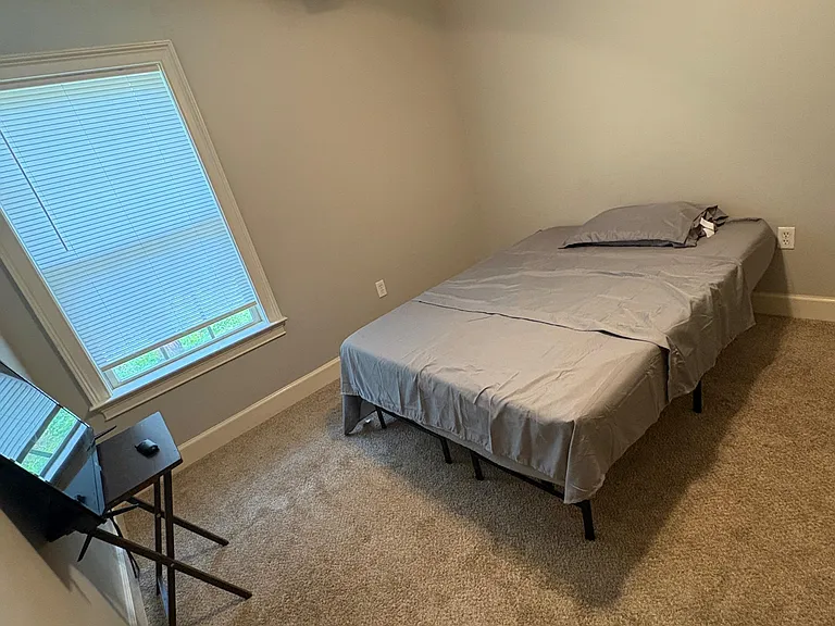 Carpeted bedroom with a ceiling fan, a large window with blinds, and space for a full bed plus a work desk.