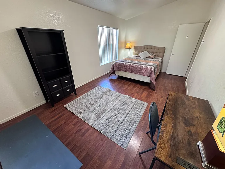 Furnished bedroom with a full bed, window with blinds, desk workspace, and storage shelving on durable wood-look floors.