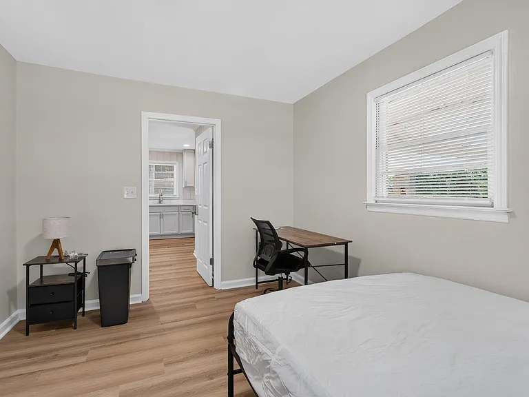 Bedroom with wood-style flooring, a large window with blinds, and space for a bed plus a dedicated work desk area.