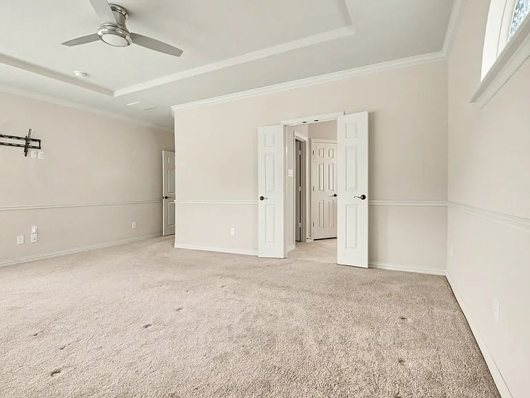 Spacious room featuring neutral carpeting, a tray ceiling with a ceiling fan, crown molding, chair rail detailing, and a double-door entry