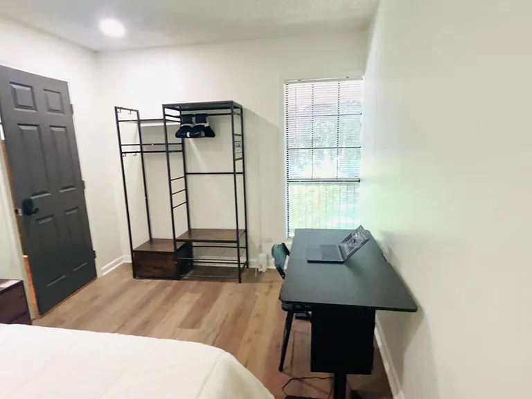 Furnished bedroom with wood-look floors, a work desk, open wardrobe storage, and a window with blinds.