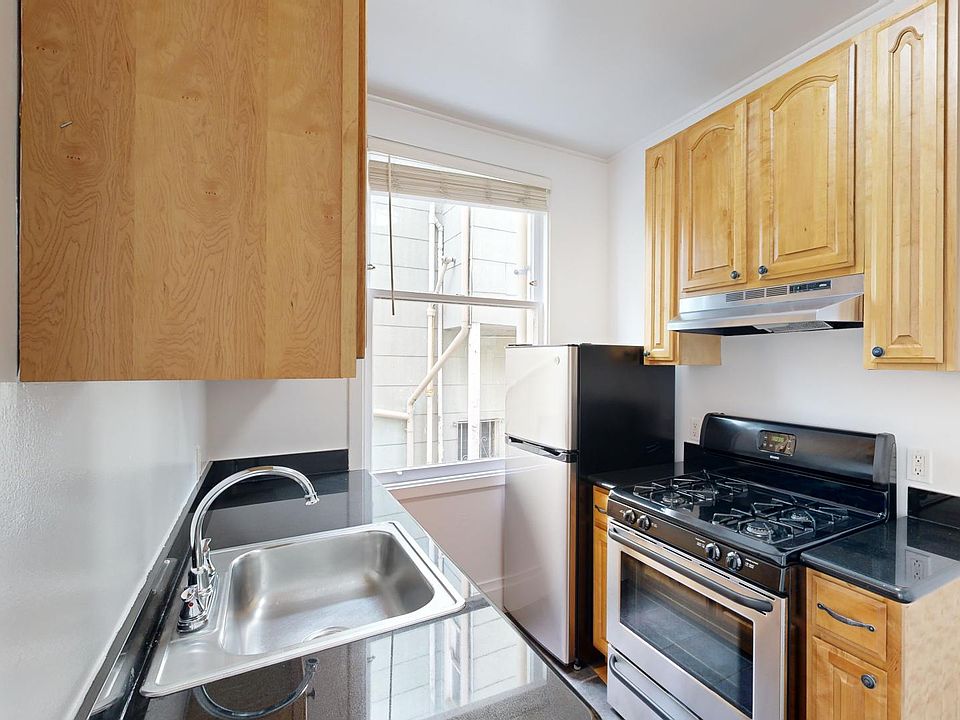 Compact kitchen with wooden cabinets, a stainless steel sink, granite countertops, a gas stove, and a small fridge. A window provides natural light.