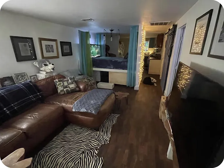 Open-concept living area with a large sectional, mounted TV space, and adjoining sleeping nook separated by curtains.