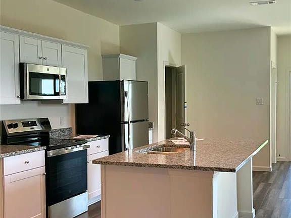 Kitchen featuring appliances with stainless steel finishes, a kitchen island with sink, light stone counters, dark wood-type flooring, and white cabinetry
