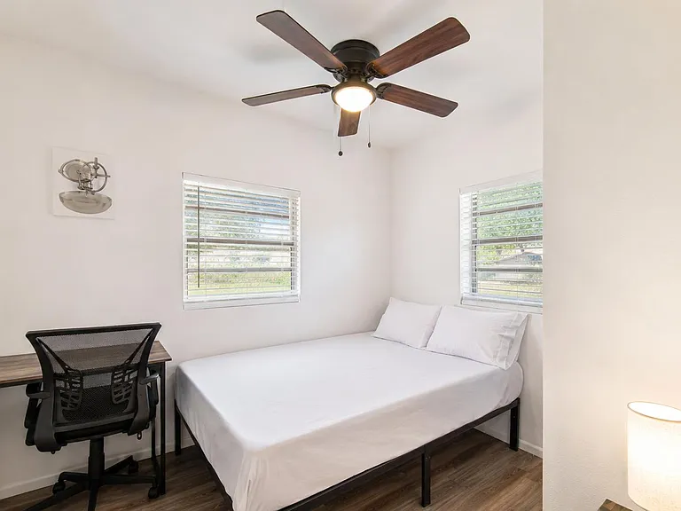 Furnished bedroom with a full bed, dedicated desk workspace, ceiling fan with light, and two windows with blinds.
