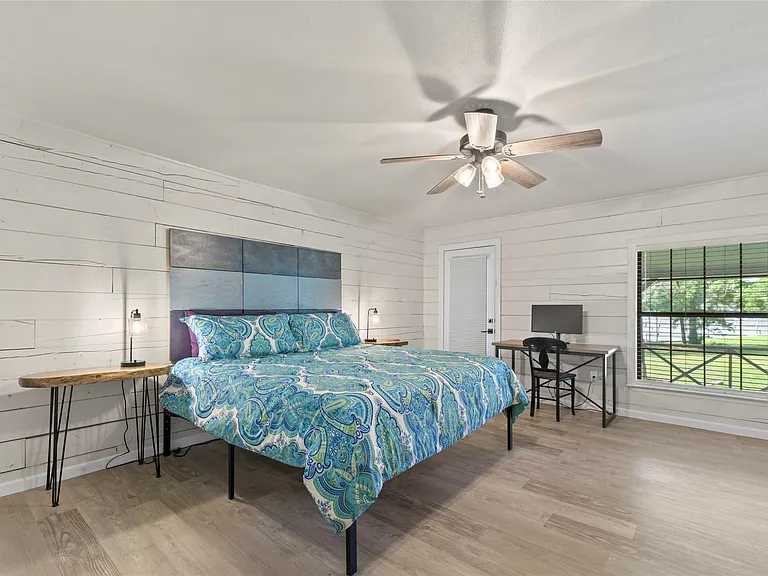 Bedroom featuring wood-finish flooring, white shiplap walls, a ceiling fan with light fixture, a window with blinds, and a white interior door
