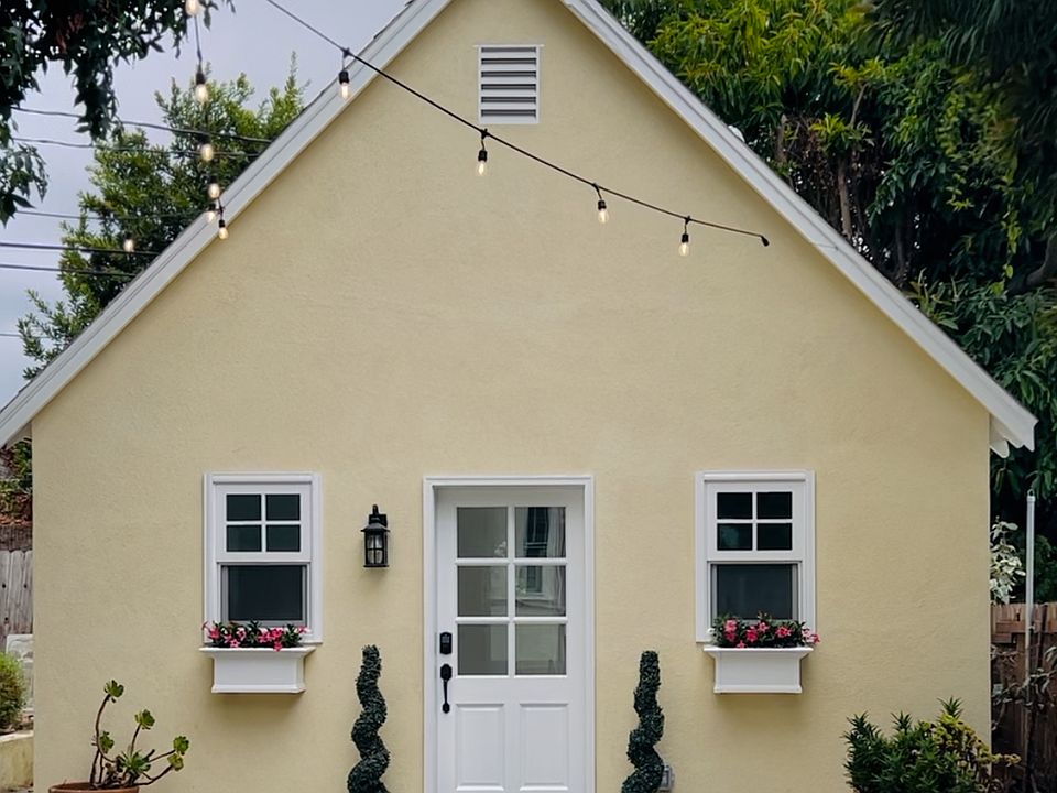 A charming new cottage with great curb appeal. Photo features a front paver patio and driveway, strung bistro lights, and two welcoming window boxes with real flowers. The classic hardwood door has a digital lock for security and convenience.
