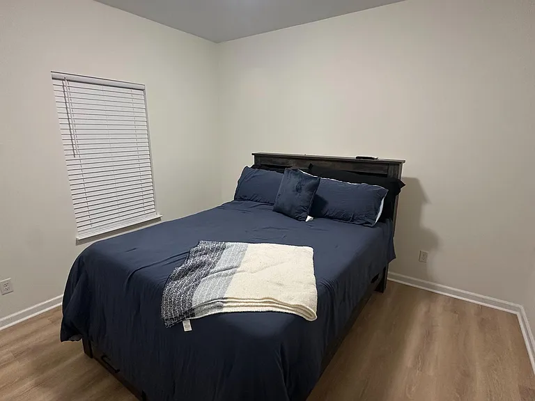 Furnished bedroom with a full bed, wood-style flooring, and a window with blinds for light control and privacy.