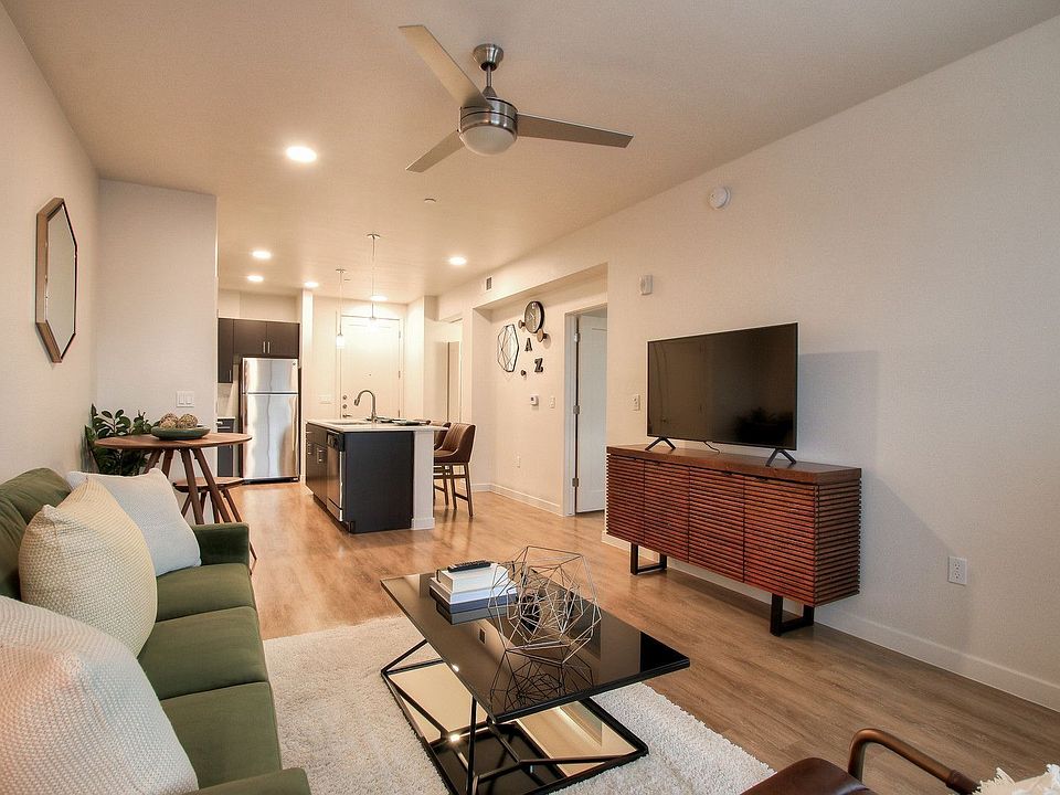 White living room with modern ceiling fan, view to kitchen with large island, and door to bedroom.