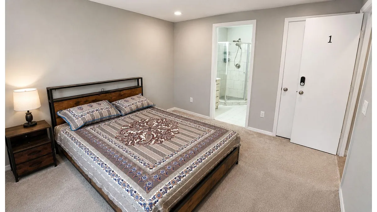 Furnished bedroom with a queen bed, nightstand, and direct access to a private bath with a glass-enclosed shower. - SW 39th Ave