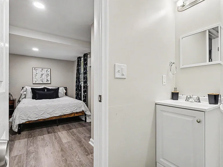 Private bedroom with a full-size bed, wood-look floors, and a window with curtains; adjacent vanity area adds convenience.