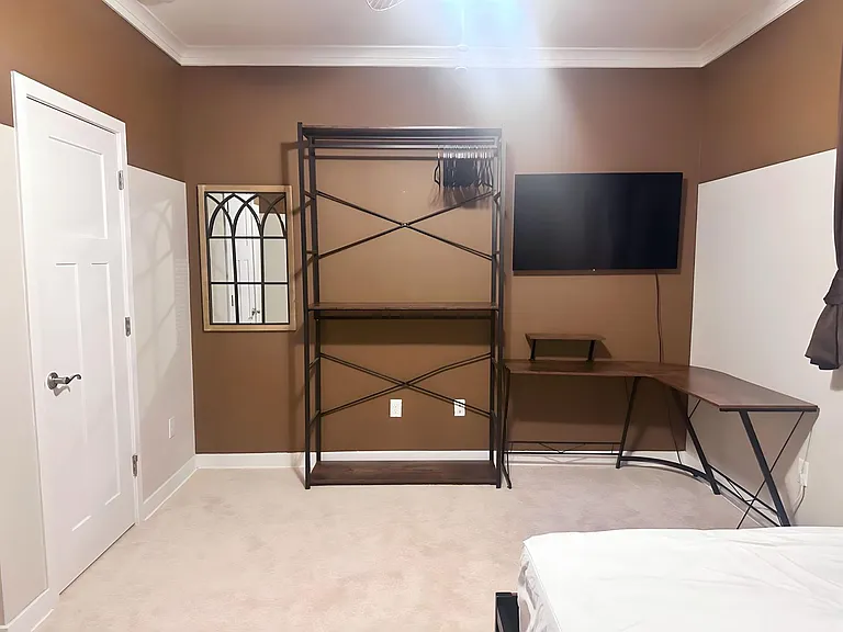 Bedroom with carpeted flooring, wall-mounted TV, corner desk setup, and open garment rack for convenient storage.