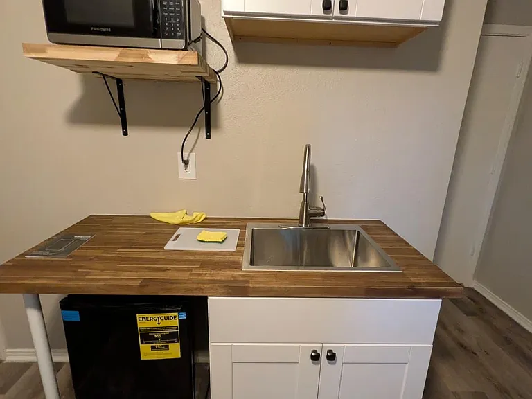 Compact kitchenette with stainless sink, wood-look countertop, upper/lower cabinetry, and included microwave and mini fridge.