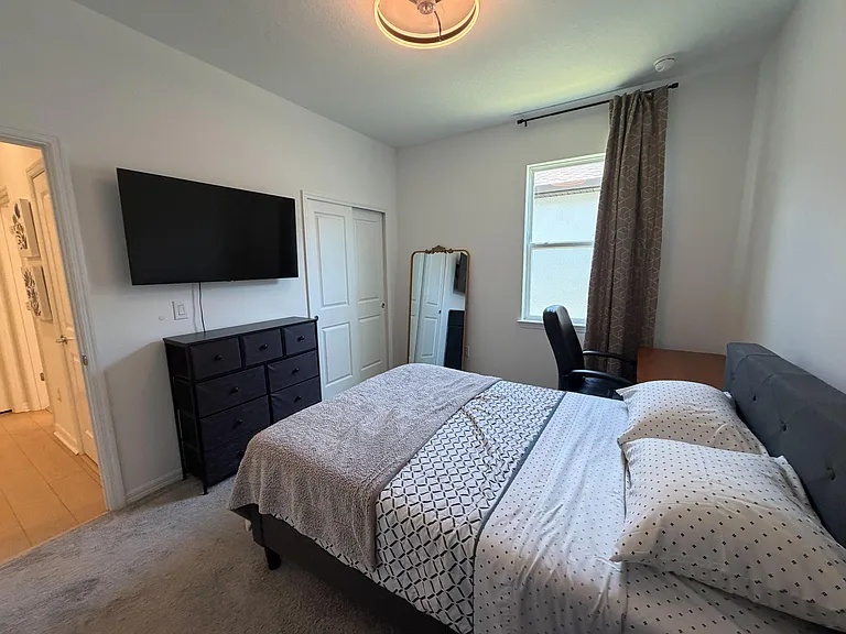 Furnished bedroom with a full bed, dresser, wall-mounted TV, and a desk area, plus a closet and window with curtains.