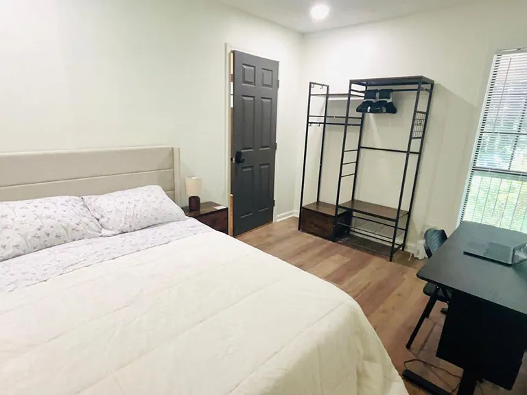 Furnished bedroom with a full bed, workspace, and open closet system, plus a large window with blinds for natural light.