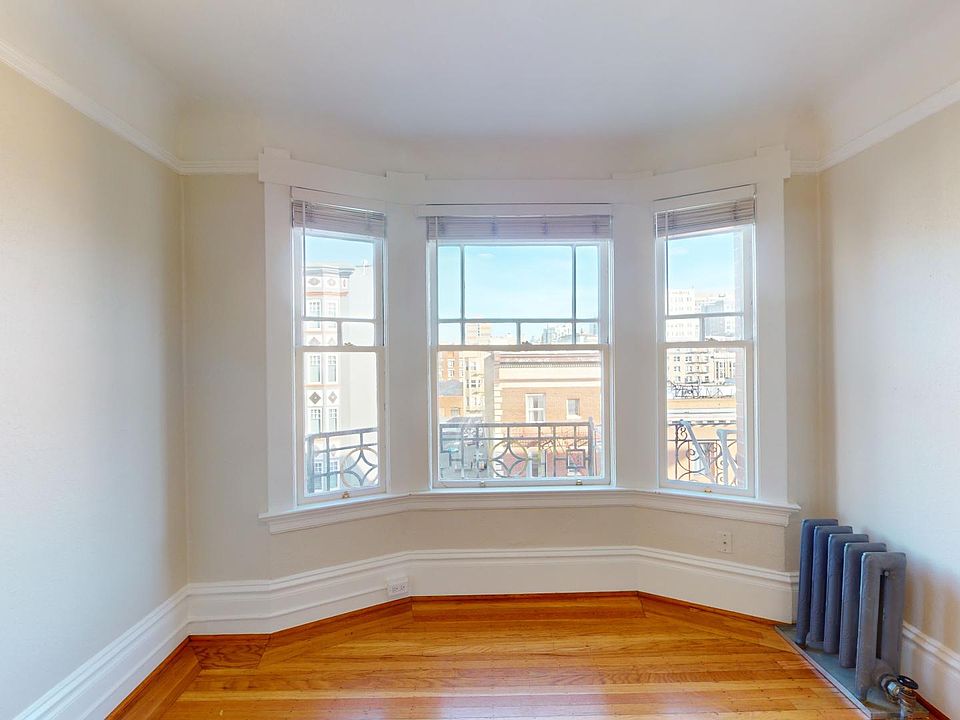 Empty room with wooden floor, walls, and a large bay window overlooking an urban landscape. A vintage radiator is in the corner, creating a cozy atmosphere.