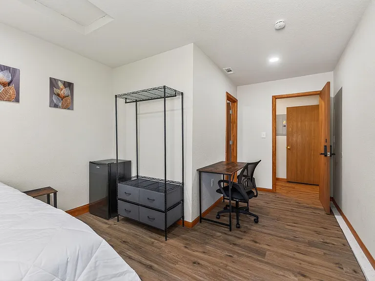 Furnished bedroom with wood-look flooring, a dedicated desk/work area, mini fridge, and open shelving for easy storage.