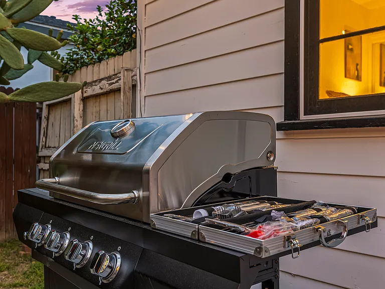 Cozy backyard retreat with cafe-style string lights, outdoor dining, a BBQ grill, and a fire pit lounge. Surrounded by palm trees and warm lighting, this inviting space is perfect for relaxing evenings and group gatherings.