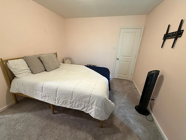 Furnished bedroom with a queen bed, soft carpeting, and a wall-mounted TV bracket, offering a simple, functional layout.