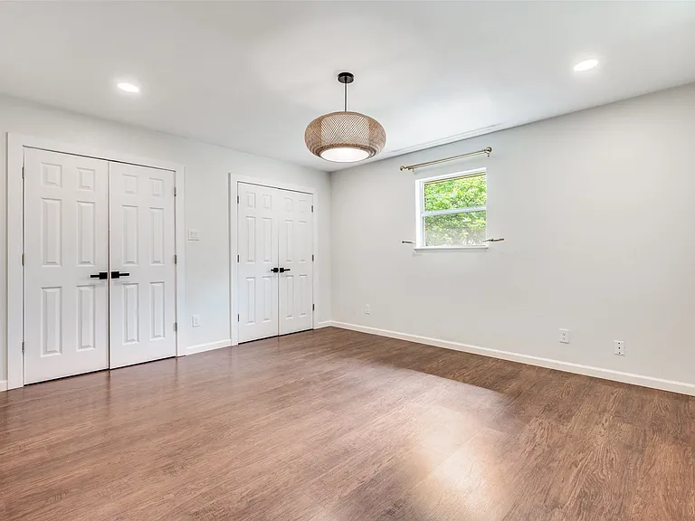 Spacious room featuring wood-finish flooring, two sets of bi-fold doors with matte black hardware, a window with exterior greenery, recessed lighting, and a drum pendant light fixture