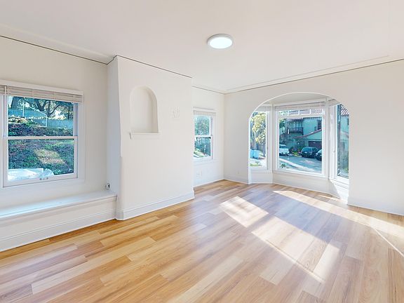 Spacious living room with a large window offering a view of a tree-lined street and with a wooden floor