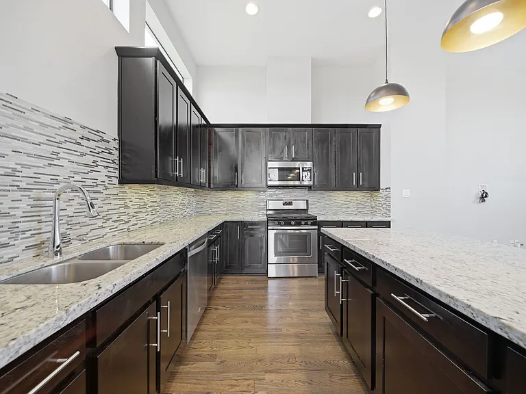 Contemporary kitchen designed to impress, featuring rich dark wood cabinetry, stainless steel appliances, and a stylish tile backsplash. The expansive layout is anchored by a large granite island with elegant pendant lighting.