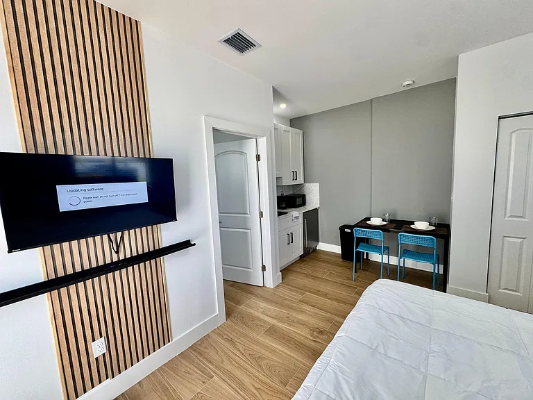 Private bedroom suite with mounted TV, wood-look flooring, and an in-room kitchenette plus a two-seat dining/work table.