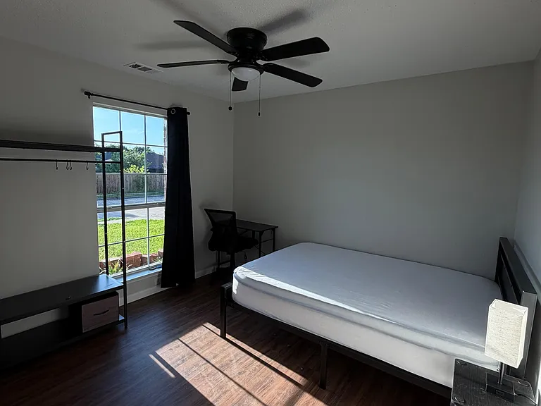 Private bedroom with wood-look flooring, ceiling fan, and a large window with curtains plus space for a desk or dresser.