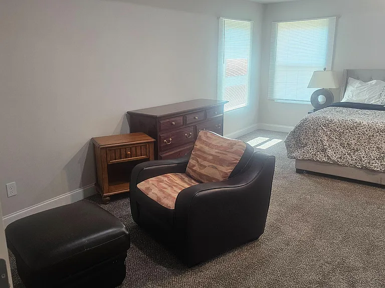 Spacious furnished bedroom with a comfortable bed, dresser storage, and a sitting area by dual windows with blinds.