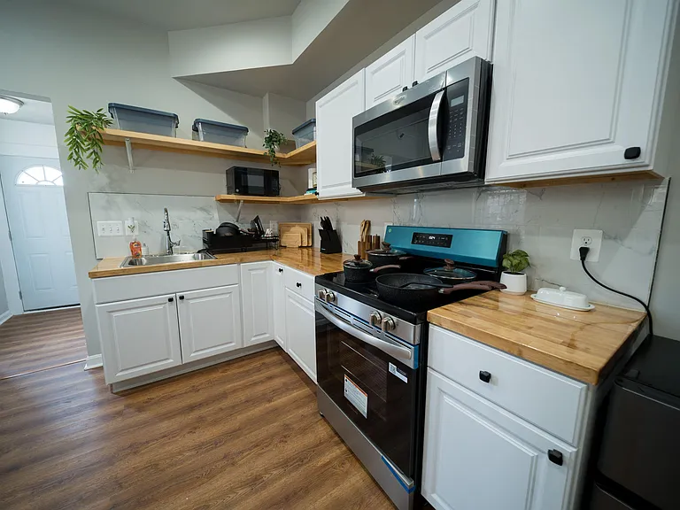 Efficient L-shaped kitchen with white cabinetry, butcher-block counters, open shelving, and stainless range with microwave.