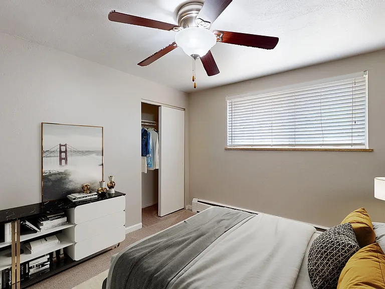 Bright bedroom with modern ceiling fan, cozy bed with stylish cushions, and ample natural light filtering through blinds.