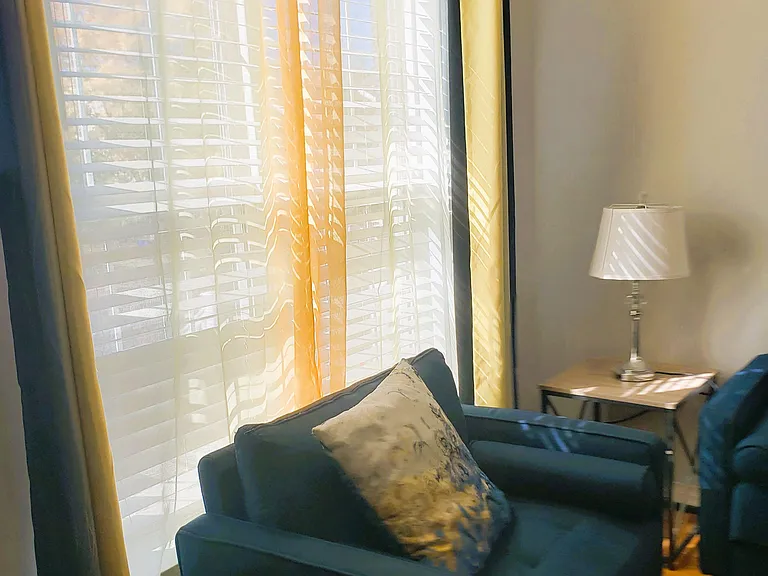 Living area with a large window, layered blinds and curtains, plus comfortable seating and a side table for everyday use.