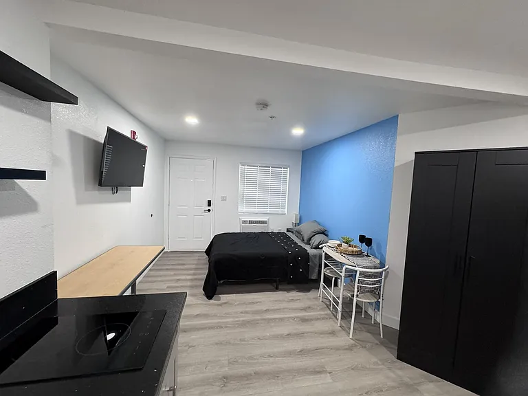 Efficient studio-style bedroom with a wardrobe, wall-mounted TV, desk space, and a window AC unit for comfort.