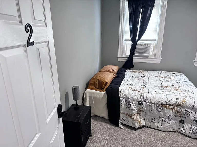 Carpeted bedroom with a full bed setup, nightstand, and window A/C unit for added comfort and climate control.