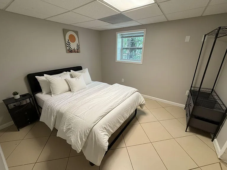 Private bedroom with a full bed, nightstand, and open shelving, plus a window and durable tile flooring.
