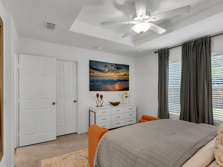 Spacious bedroom with tray ceiling and ceiling fan, large window with blinds and curtains, and a dresser for storage.