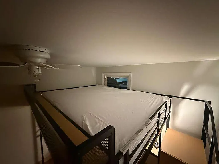 Compact loft-style sleeping area with a metal bed frame, ceiling fan, and small window for light and ventilation.