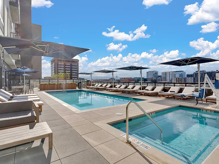 Luxurious rooftop pool area with modern lounge chairs, stylish umbrellas, and stunning cityscape views under a bright blue sky.