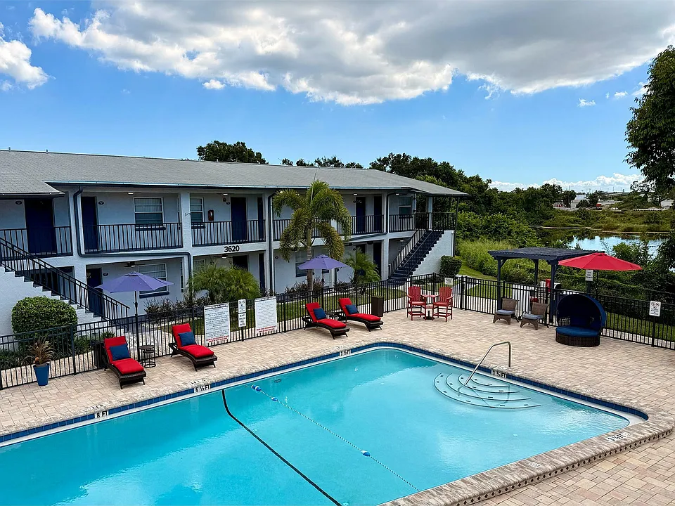 Charming apartment complex featuring a picturesque pool area with red lounge chairs, umbrellas, and lush greenery backdrop.