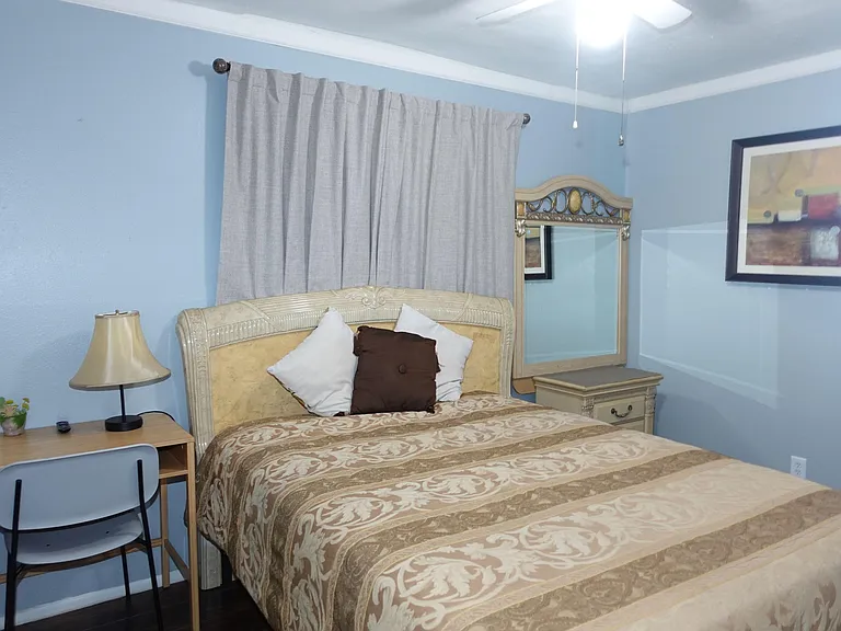 Furnished bedroom featuring a queen bed, nightstand and lamp, plus a small desk area and window with curtains.