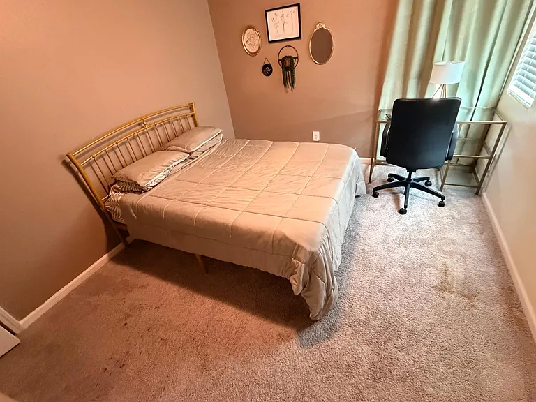 Furnished bedroom with a full bed, soft carpet, and a dedicated desk setup near the window for work or study.
