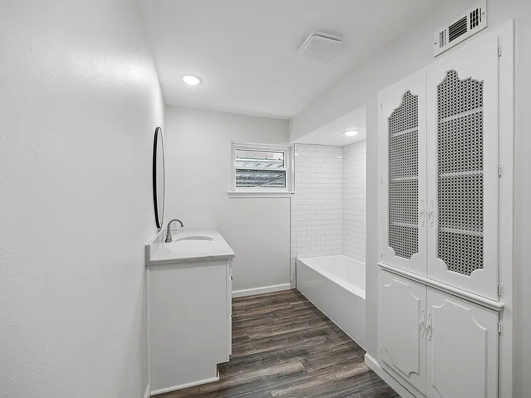Bathroom featuring a white vanity with a rectangular sink and stone-finish countertop, a tub with subway tile surround, wood-finish flooring, and a built-in storage cabinet