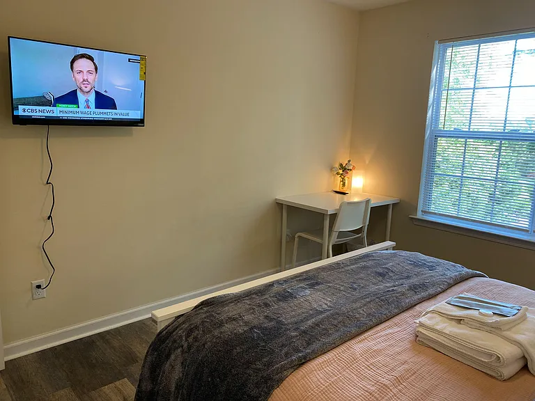 Bedroom includes a wall-mounted TV, a dedicated desk nook, and a large window with blinds for natural light.