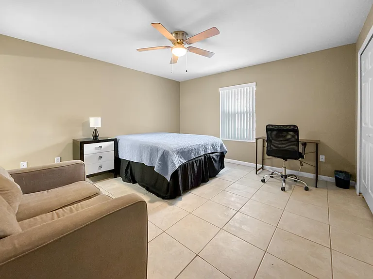 Spacious furnished bedroom with tile flooring, ceiling fan, and a window with blinds plus a desk area for work or study.