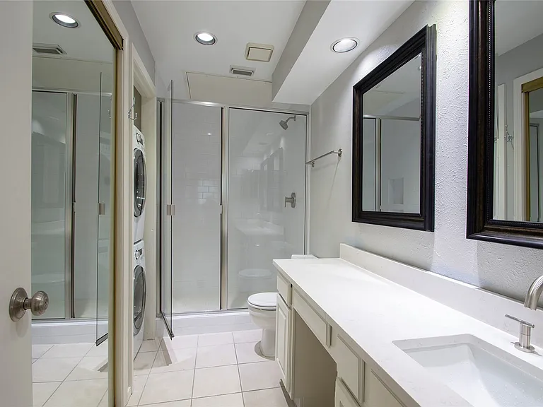 White tile flooring in a bathroom featuring a long vanity with an integrated sink and brushed nickel faucet, framed mirrors, a glass-enclosed shower, and recessed lighting
