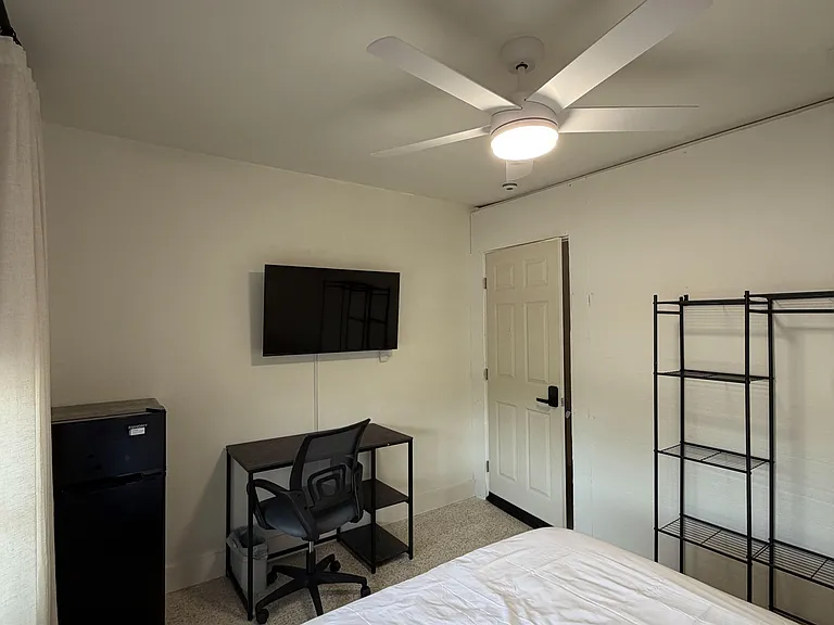 Private bedroom with a ceiling fan, mounted TV, mini fridge, work desk, and open shelving for easy storage.