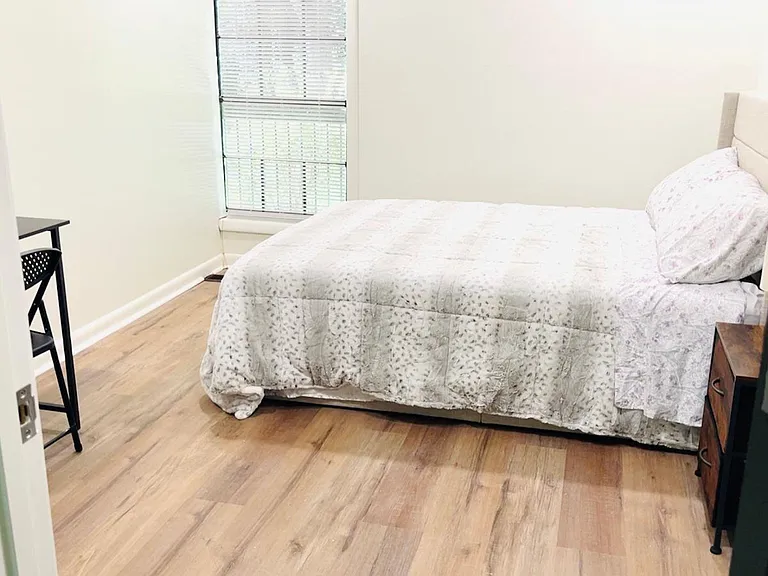 Private bedroom with a full bed, bedside table, and a small desk area, plus a window with blinds and wood-style flooring.