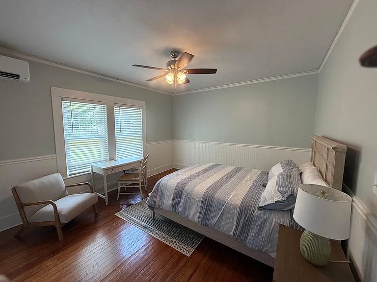 Furnished bedroom with hardwood floors, ceiling fan, and large windows, plus a desk nook for work or study.