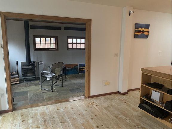 From dining room looking toward stone-floor living room. There are large wooden garage doors doors that open to a garden area to the left of the wood-fired stove.